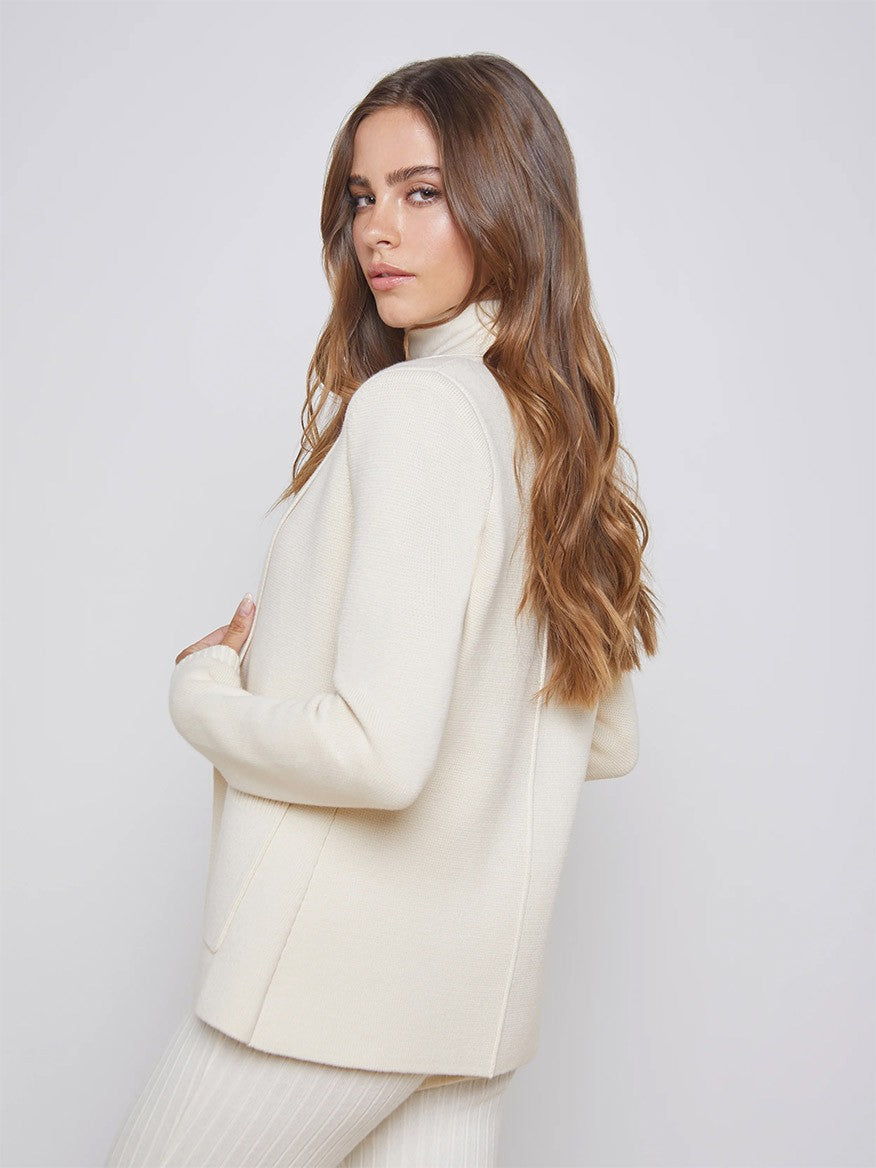 A woman with long, wavy hair wearing a L'Agence Lacey Knit Blazer in Porcelain, posing with her back partially turned and looking over her shoulder.