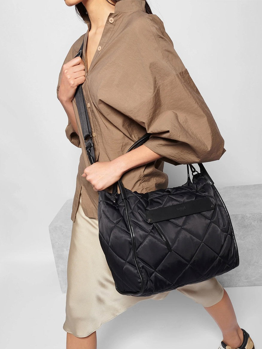 A person in a beige jacket carrying a large black MZ Wallace Quilted Large Madison Shopper in Black Bedford.