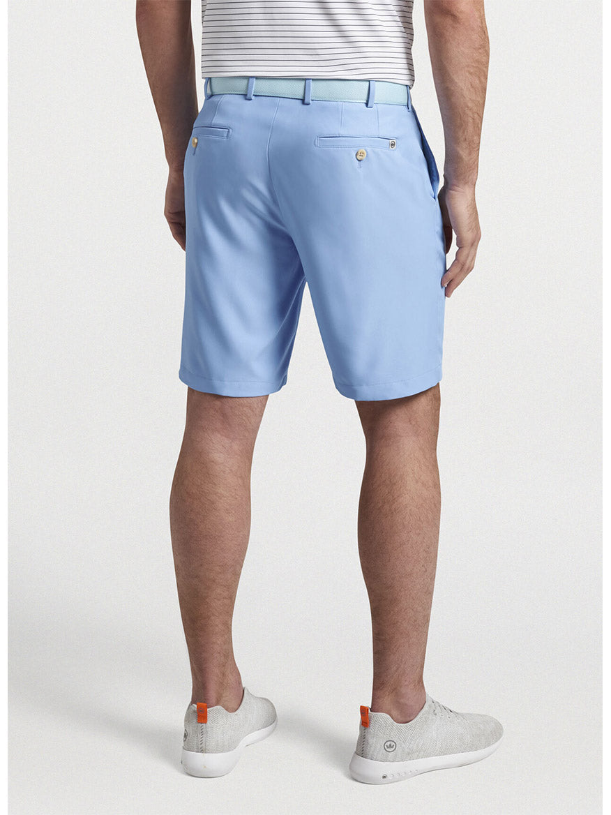 Man standing in profile wearing the Peter Millar Salem High Drape Performance Short in Cottage Blue, a striped belt, white shirt, and gray sneakers.