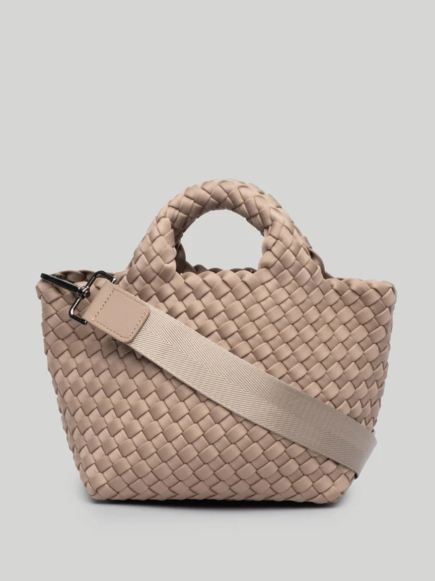 The Naghedi St. Barths Petit Tote in Solid Cashmere, a beige woven handbag with two short handles and a long crossbody strap, is set against a light gray background.