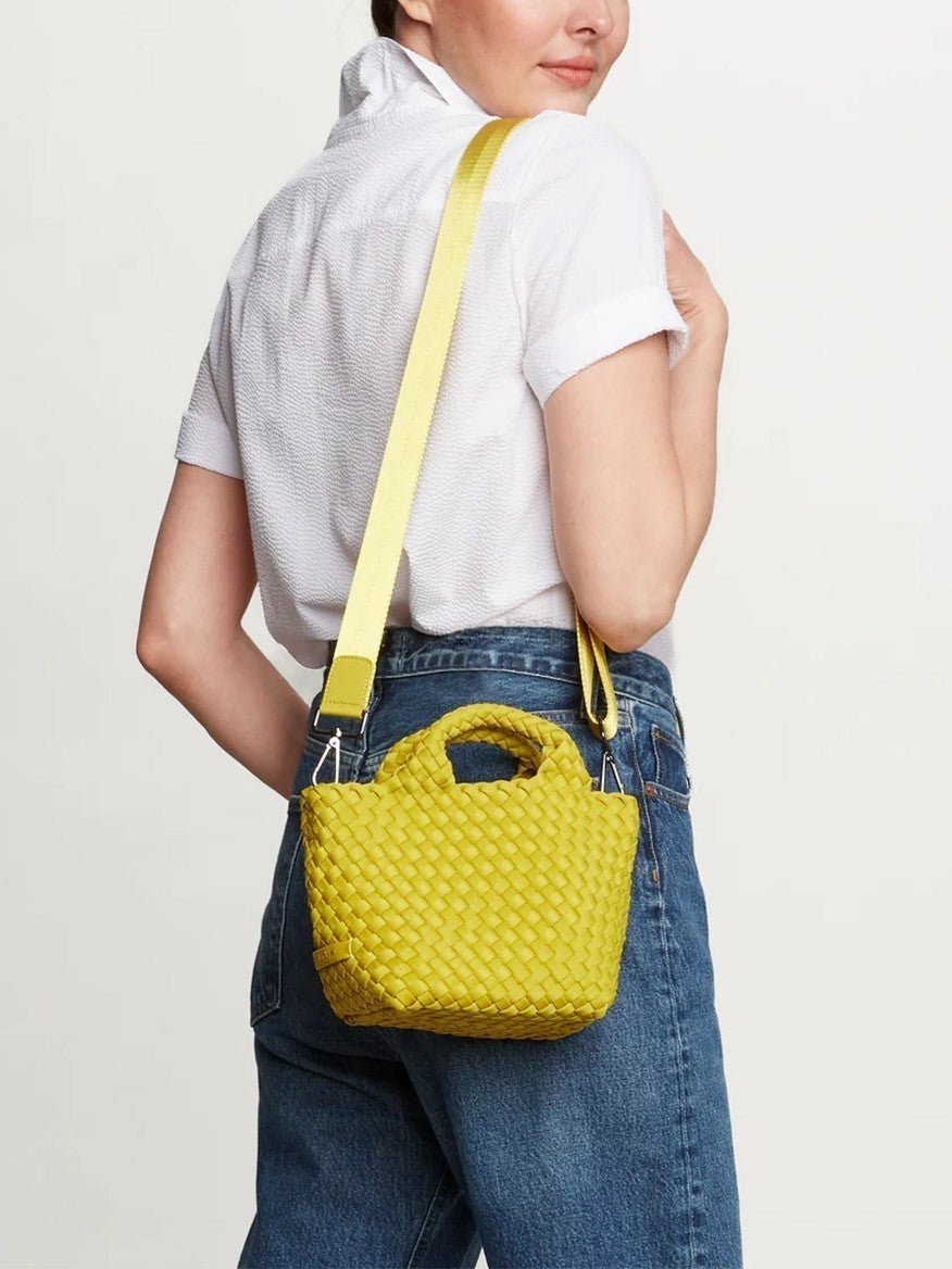 A person in a white shirt and blue jeans is seen from the back, carrying a yellow Naghedi St. Barths Petit Tote in Solid Cashmere with a crossbody strap.