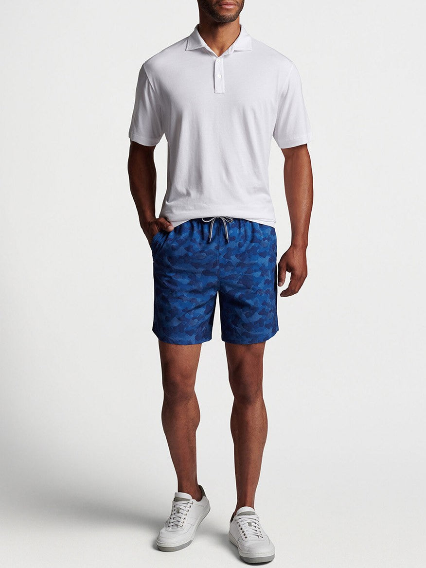 Man wearing a white polo shirt, blue patterned Peter Millar Camouflaged Coastline Swim Trunk in Navy with a 7" inseam, and white sneakers.