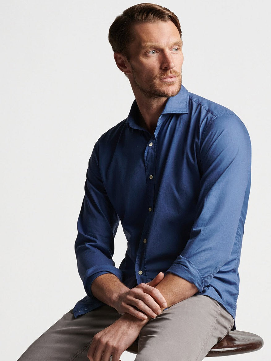 Man in a Peter Millar Sojourn Garment-Dyed Cotton Sport Shirt in Blue Pearl and gray pants sitting on a stool, looking to the side with a thoughtful expression.