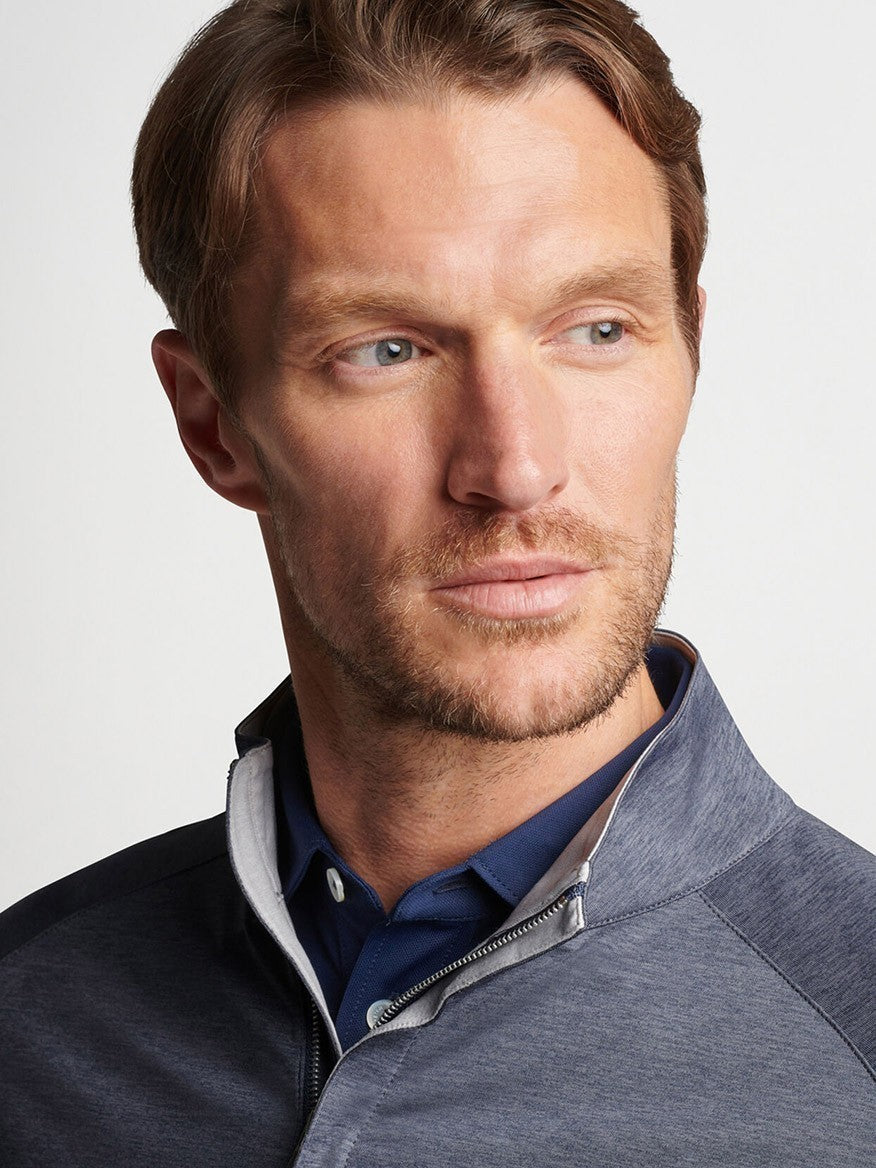 A man with stubble wearing a blue Peter Millar Stealth Performance Quarter-Zip in Steel shirt and a gray quarter-zip sweater, looking to the side.
