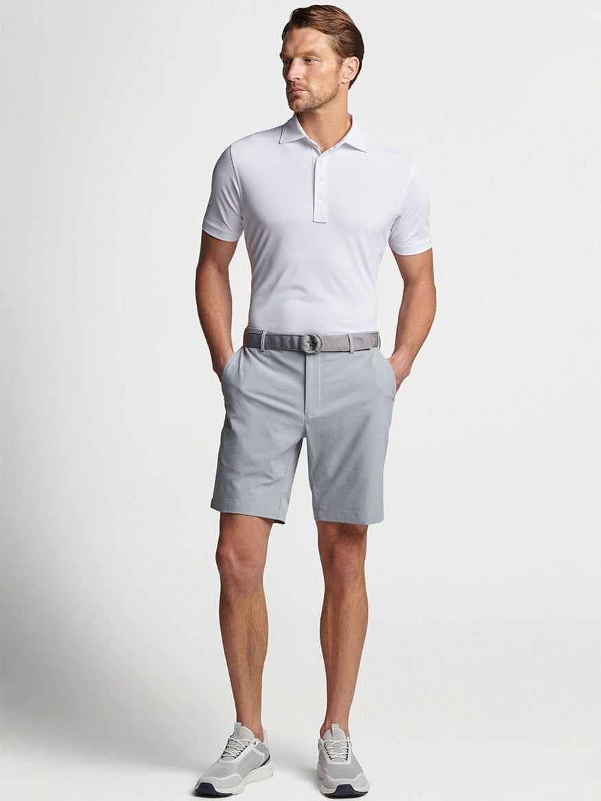 A man modeling a casual light grey Peter Millar Surge Performance Short in Gale Grey and white Peter Millar polo shirt outfit, with a belt and sneakers, showcasing performance apparel.