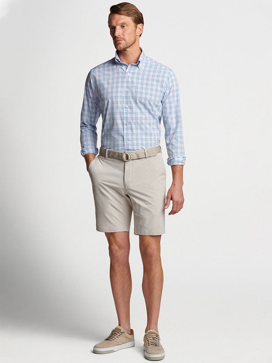 Man modeling a casual summer outfit featuring a blue checkered Peter Millar shirt, [Peter Millar Surge Performance Short in Oatmeal], and sneakers designed for water-resistant protection.