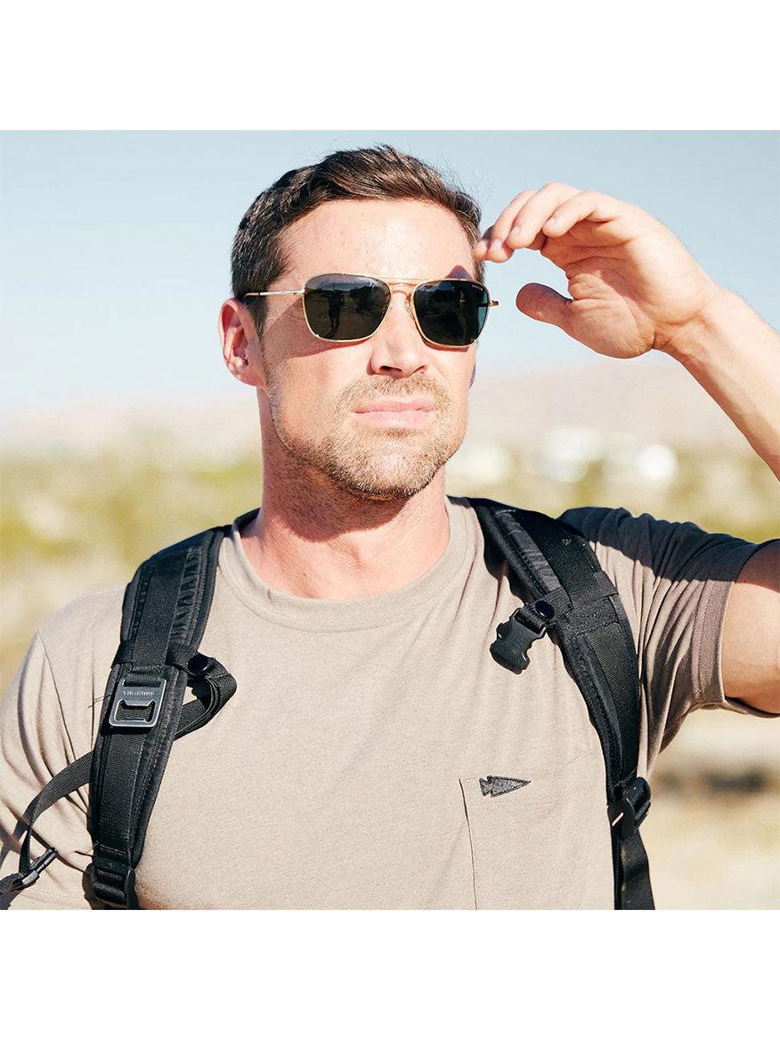 A man wearing Randolph Intruder American Gray Sunglasses in Matte Black and carrying a backpack stands outdoors in a sunny setting, savoring the clarity of the lenses.