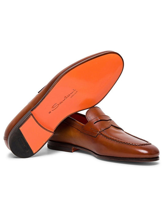 A pair of Santoni Door Leather Penny Loafers in Light Brown with orange soles, one shoe lying flat and the other propped against it, showcasing the brand name "Santoni" on the sole. This classic loafer is crafted from premium quality leather and features meticulous Goodyear construction.