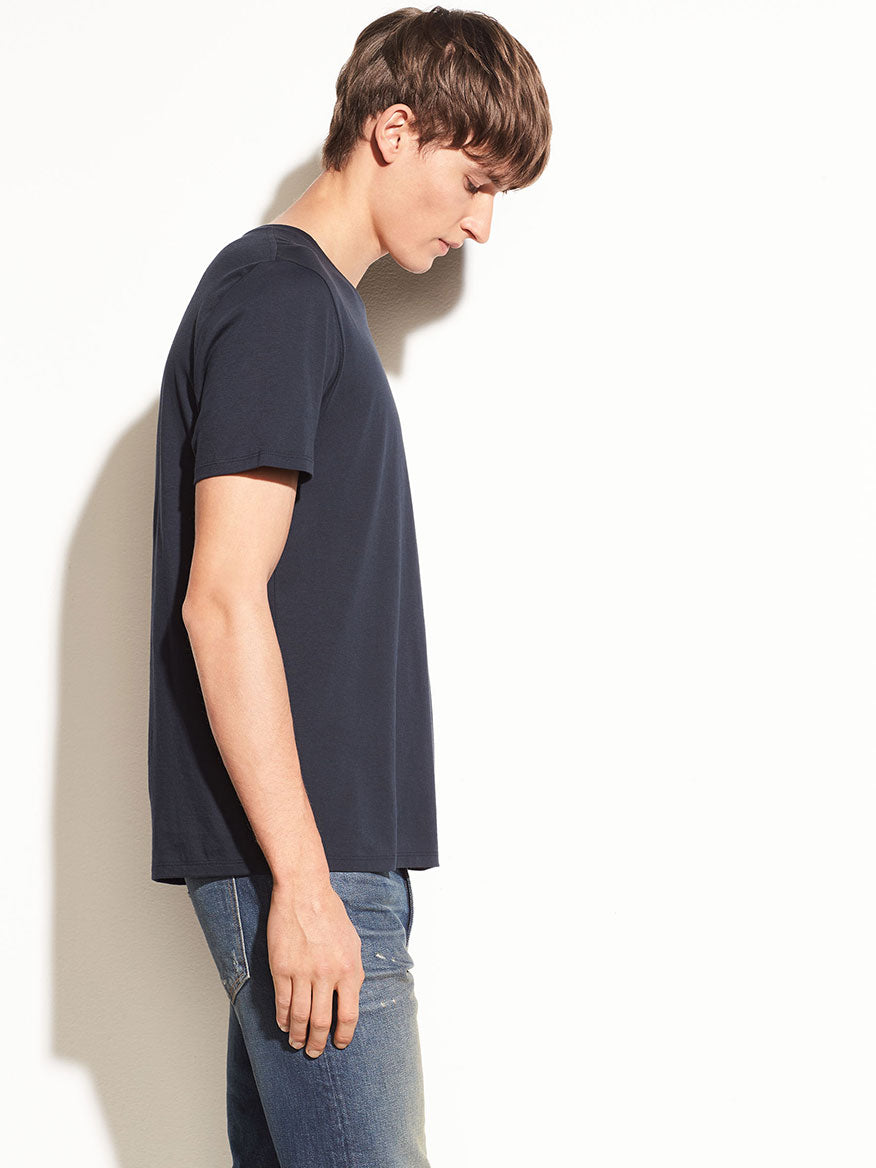 Young man in a Vince Crew Neck T-Shirt in Coastal Blue and jeans standing against a light wall, looking downward with a neutral expression.