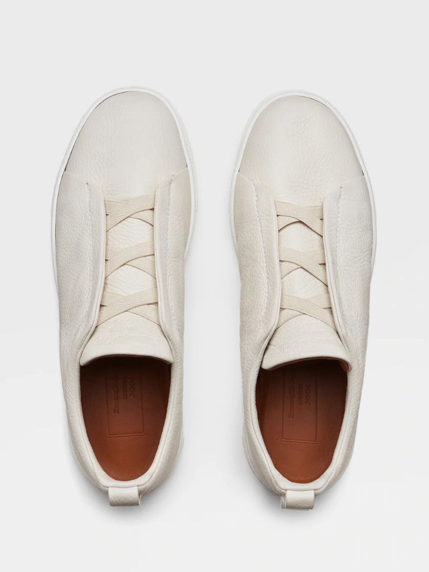 A top-down view of a pair of Zegna Off White Deerskin Triple Stitch™ Sneakers with intricate lace design and brown insoles, displayed on a white background.